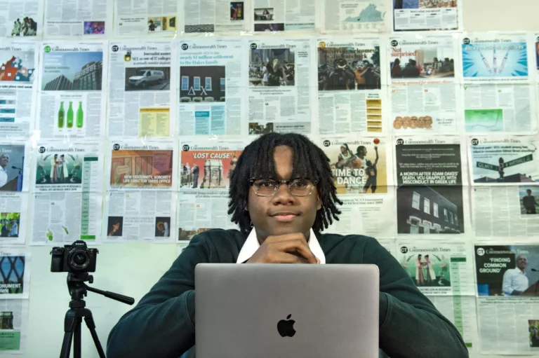 A man sitting in front of an Apple laptop with his hands under his chin. Behind him are layouts of newspaper pages.